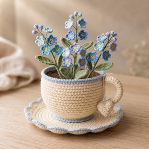 Könnte beinhalten: Ein gehäkeltes cremefarbenes Tassen- und Untertassenset mit hellblauem Rand, das einen Strauß aus gehäkelten blauen Vergissmeinnicht-Blumen mit grünen Blättern enthält. Ein kleiner gehäkelter Herzanhänger hängt am Henkel der Tasse.