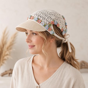 May include: A cream-colored crochet baseball cap featuring a solid woven brim and a decorative band of small, multi-colored fabric flowers in shades of blue, peach, and green. The cap includes a tie-back closure with matching crochet ribbons.