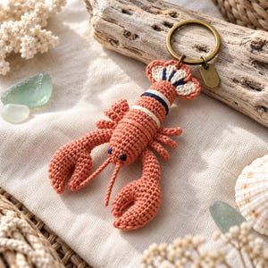 Può includere: Un portachiavi a forma di aragosta all'uncinetto con corpo color corallo, strisce blu scuro e bianche, due grandi chele e due antenne. L'aragosta è fissata a un anello metallico color bronzo con una piccola targhetta rettangolare.