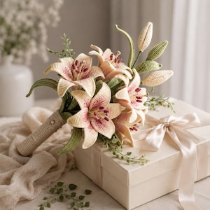May include: A bouquet of crocheted lily flowers in shades of cream and soft pink with dark pink speckles. The flowers feature green crocheted leaves and stems, with a wrapped handle. The bouquet is positioned next to a cream-colored gift box.