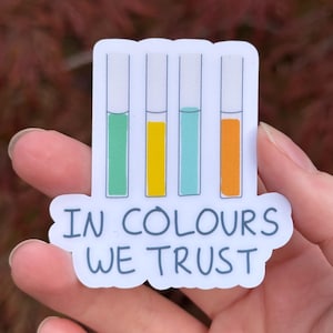 Peut inclure: Un autocollant découpé représentant quatre tubes à essai verticaux remplis de liquide vert, jaune, bleu clair et orange. Sous les tubes, le texte "IN COLOURS WE TRUST" est imprimé en bleu foncé, police sans empattement sur fond blanc.