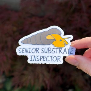 May include: A die-cut sticker featuring a cartoon snail with a grey shell and a bright yellow body. The text SENIOR SUBSTRATE INSPECTOR is printed in blue capital letters below the snail illustration against a white background.