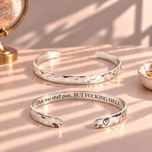May include: Two silver-toned cuff bracelets with a hammered, textured exterior. The interior of one bracelet is engraved with the text "This too shall pass, BUT FUCKING HELL" and features a small black heart symbol near the open end of the band.