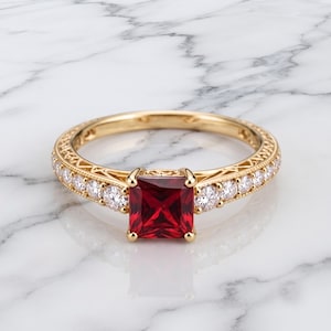 May include: A gold ring featuring a square-cut deep red gemstone at the center, flanked by a row of small round clear stones on each side. The gold band displays intricate filigree metalwork along the sides, providing a detailed texture.