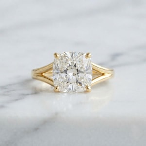 May include: A gold ring featuring a large, clear, cushion-cut gemstone set in a four-prong mounting. The band is crafted from polished yellow gold and splits into two distinct, slender arms as it reaches the central stone, creating a bypass design.