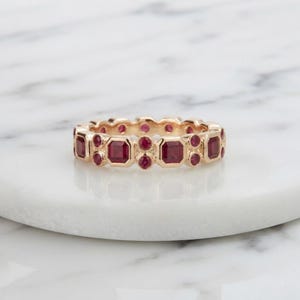 Peut inclure: Une bague en or rose ornée d'un motif alterné de rubis rouge foncé taille carrée et taille ronde, sertis en une rangée continue sur toute la circonférence de l'anneau métallique, formant un bijou symétrique et détaillé.