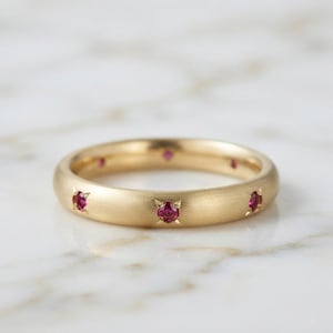 May include: A gold band ring featuring a brushed matte finish and multiple small, round, vibrant red rubies set flush into the metal at regular intervals around the circumference of the circular jewellery piece.