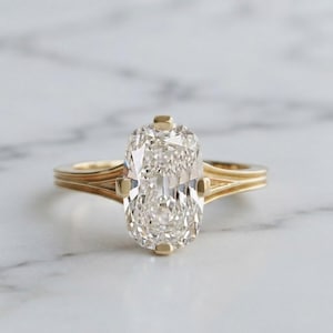 May include: A gold ring featuring a large, elongated cushion-cut clear gemstone set in a four-prong setting. The band is crafted from yellow gold and displays a distinct triple-ribbed or grooved design that extends from the shoulders to the shank.