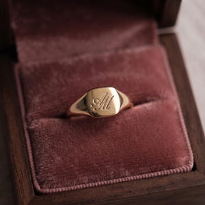 Könnte beinhalten: Ein goldener Siegelring mit einer glatten, polierten Oberfläche und den eingravierten Schreibschriftbuchstaben AM in der Mitte. Der Ring hat eine sich verjüngende Ringschiene und einen abgerundeten quadratischen Kopf mit metallischem Glanz.
