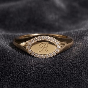 May include: A gold signet ring featuring an oval face with a polished center engraved with the letter R. The oval face is surrounded by a halo of small, clear, round-cut stones set into the gold band, which has a smooth, reflective finish.