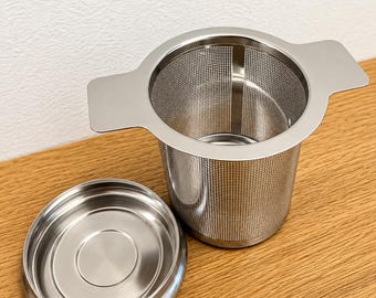 May include: A stainless steel tea infuser basket with a fine mesh body and two flat side handles, accompanied by a matching circular lid. The metallic silver accessory features a cylindrical shape designed for steeping loose leaf tea in a mug.