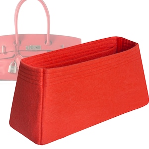 May include: A bright red felt handbag organizer with a rectangular shape. The organizer has multiple compartments and is designed to fit inside a handbag. The background shows a blurred image of a pink handbag.