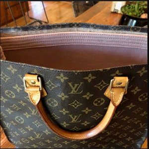 May include: A brown and tan leather tote bag with a brown and tan monogram pattern. The bag has a pink fabric lining and two brown leather handles.