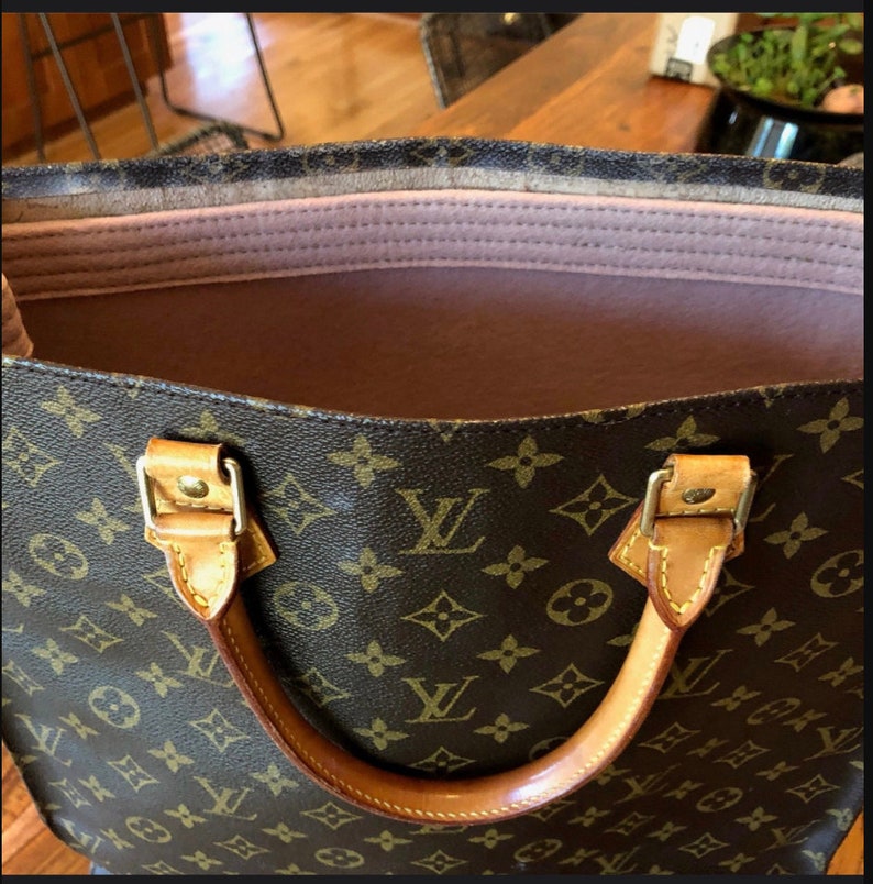 May include: A brown and tan leather tote bag with a brown and tan monogram pattern. The bag has a pink fabric lining and two brown leather handles.