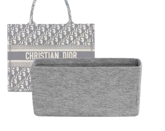 Puede incluir: Un inserto para bolso tote de fieltro gris con forro de tela gris. El inserto está diseñado para caber dentro de un bolso tote con un patrón blanco y gris y el texto "CHRISTIAN DIOR" impreso en la parte delantera.