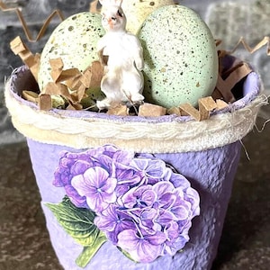 May include: A purple textured pot decorated with a purple hydrangea flower cutout and a white braided trim. Inside the pot are three speckled light green eggs, a small white rabbit figurine, and brown paper filler strips.