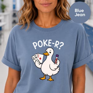 May include: A blue jean coloured t-shirt featuring a graphic of a white duck holding three playing cards and a medical blood collection tube. Above the duck, the white text reads POKE-R? The shirt has short sleeves with rolled cuffs.