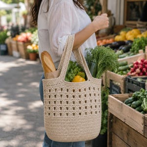 May include: A beige crochet tote bag with a structured base and an open-work grid pattern on the upper section. The bag features two sturdy handles and is filled with a baguette, a yellow bell pepper, green lettuce, and fresh herbs.