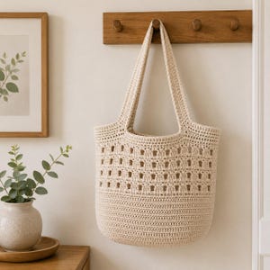 May include: A cream-colored crochet tote bag featuring a solid base and a top section with a repeating open-work square pattern. The bag has two long shoulder straps and is constructed from a thick, textured yarn in a neutral off-white shade.