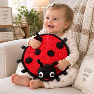 Peut inclure: Un jouet coccinelle rond en crochet rouge avec des points noirs, une tête noire et des yeux blancs. Le jouet présente de petites boucles en tissu noir fixées sur le pourtour du corps circulaire, offrant des éléments tactiles pour le jeu.
