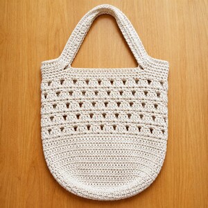 May include: A cream-colored crochet tote bag featuring a dense, solid base and a top section with an open, repeating geometric stitch pattern. The bag includes a single integrated handle at the top, crafted from the same off-white yarn.