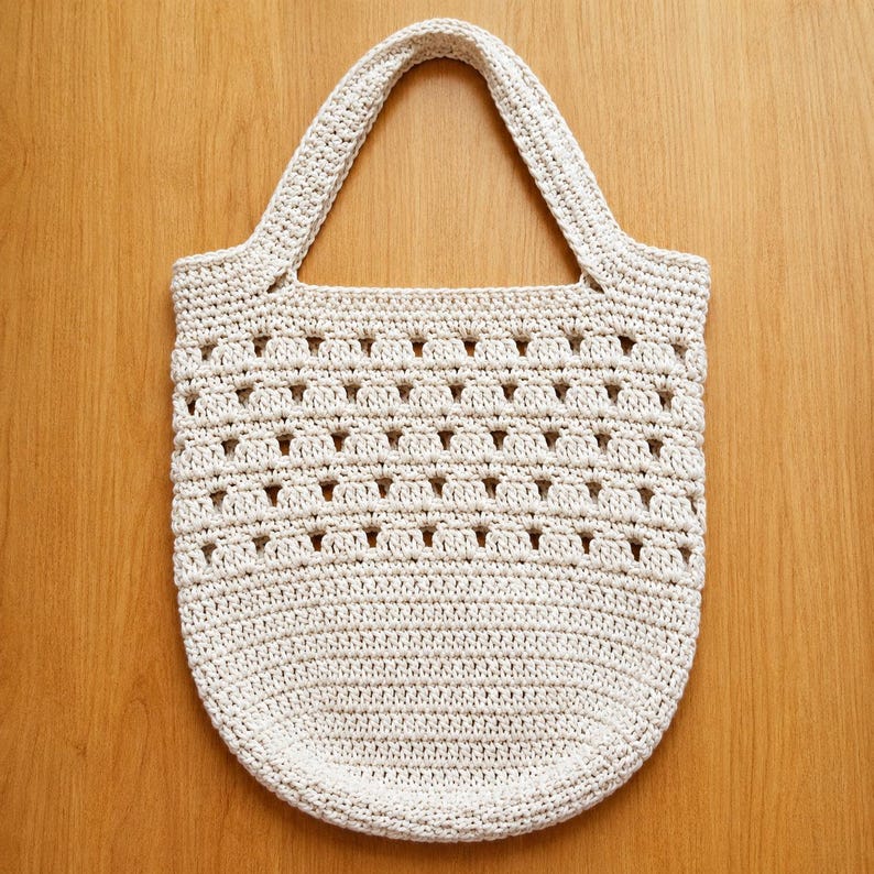May include: A cream-colored crochet tote bag featuring a dense, solid base and a top section with an open, repeating geometric stitch pattern. The bag includes a single integrated handle at the top, crafted from the same off-white yarn.