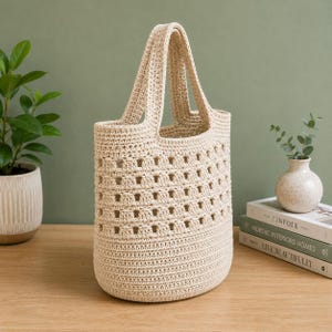 May include: A cream-colored crochet tote bag featuring a solid base and a structured upper section with a repeating square cutout pattern. The bag has two integrated handles and is crafted from a thick, durable cord material with a textured finish.