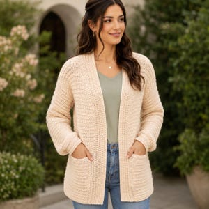 Puede incluir: Un cárdigan de punto en color beige claro abierto por delante, con mangas largas, textura acanalada y dos grandes bolsillos de parche en la parte frontal. La prenda se lleva sobre una camiseta verde salvia con cuello en V y vaqueros azules.
