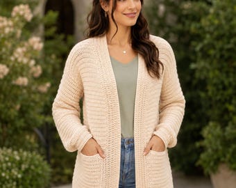 May include: A light beige, open-front knit cardigan featuring long sleeves, a ribbed texture, and two large patch pockets on the front. The garment is worn over a sage green v-neck top and blue denim jeans, showcasing a simple, functional design.