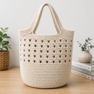 May include: A cream-colored crochet tote bag featuring a solid base and a top section with a repeating open-work square pattern. The bag has two integrated handles and is constructed from a thick, textured yarn in a neutral off-white shade.