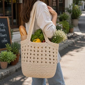 May include: A beige crochet tote bag with an openwork square pattern, featuring two shoulder straps. The bag is filled with a baguette in a paper sleeve, a yellow bell pepper, green lettuce, and a bunch of white chamomile flowers.