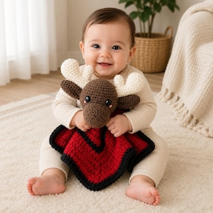 May include: A crochet security blanket featuring a brown moose head with cream-colored antlers and black safety eyes. The attached blanket is a square shape with a textured red center, bordered by a thick black crocheted edge trim.