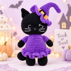 Può includere: Una bambola gatto all'uncinetto nera che indossa un vestito viola e un cappello da strega viola abbinato con una fascia arancione. Il gatto ha grandi occhi viola, baffi ricamati bianchi e un piccolo segno a x sopra un occhio.