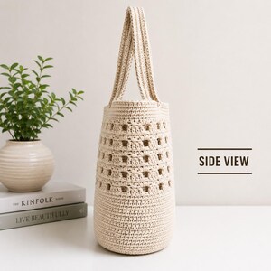 May include: A beige crochet tote bag with a tall, cylindrical shape and a single integrated handle. The bag features a dense, solid base and a middle section with an open, square-patterned weave. The text "SIDE VIEW" is printed to the right.