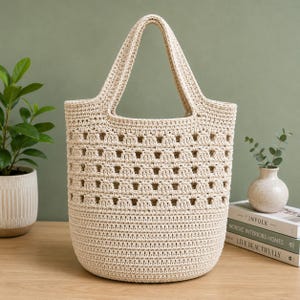 May include: A cream-colored crochet tote bag featuring a solid base and a repeating openwork pattern across the upper section. The bag has two integrated handles and a structured, rounded shape, crafted from a textured, woven cord material.