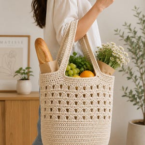 May include: A cream-colored crochet tote bag featuring a structured base and an open-work grid pattern on the upper section. The bag contains a baguette in a paper sleeve, green grapes, an orange, and a bouquet of white daisies in a paper wrap.