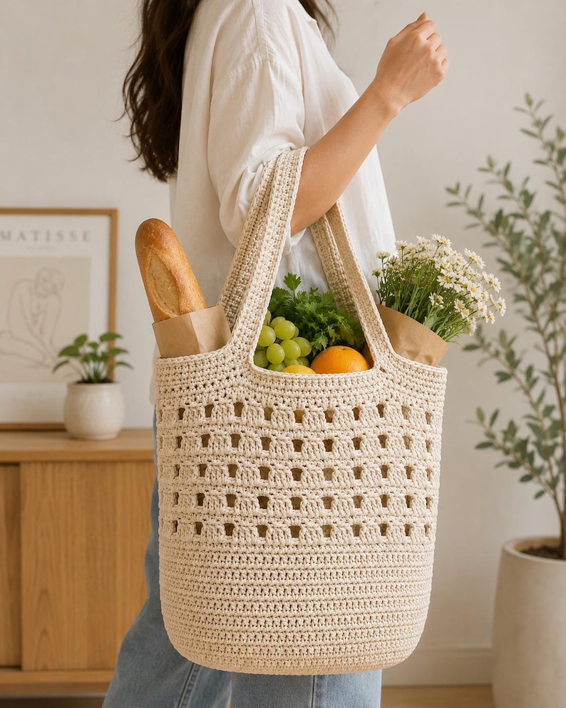 May include: A cream-colored crochet tote bag featuring a structured base and an open-work grid pattern on the upper section. The bag contains a baguette in a paper sleeve, green grapes, an orange, and a bouquet of white daisies in a paper wrap.