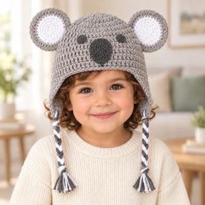 May include: A gray crocheted hat featuring a koala design with large round ears, a dark gray nose, and two braided tassels hanging from the sides. The hat is constructed with a textured stitch pattern and includes white accents on the ears.