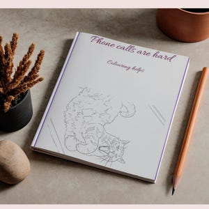 May include: A white coloring book with a purple border featuring a line drawing of a cat on the cover. The text on the cover reads "Phone calls are hard" and "Colouring helps" in purple script. The book is positioned on a flat surface.