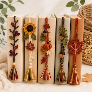 Può includere: Cinque segnalibri verticali all'uncinetto con motivi autunnali, tra cui un ramo di bacche, un girasole con foglia d'acero, una zucca con fogliame, foglie di quercia con ghiande e una grande foglia d'acero, rifiniti con perla e nappa.