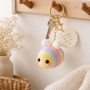 Op de afbeelding: Een gehaakte bijen-sleutelhanger met een regenbooggestreept lichaam, witte vleugels, zwarte ogen en roze wangen. Het bevat een goudkleurige metalen ring, een zilveren sleutel en een geschulpt label met de tekst: Bee happy, Bee kind, Bee you, en een geel hartje.