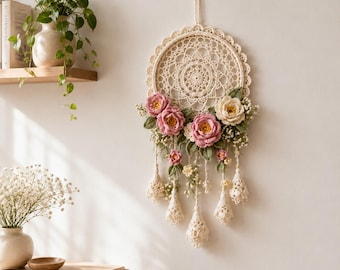 Peut inclure: Une décoration murale circulaire composée d'un napperon en crochet crème au motif complexe. La partie inférieure est ornée de fleurs en crochet roses et crème, de feuilles vertes et de petites fleurs blanches, avec des pompons en crochet.