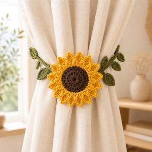 May include: A crochet sunflower curtain tieback featuring a dark brown center with bright yellow petals and two green leafy vines extending from the sides, secured around a plain off-white fabric curtain panel to hold it in place.