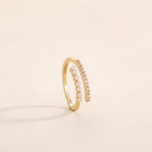 Puede incluir: Un anillo abierto de tono dorado con dos extremos paralelos. Un extremo está adornado con una hilera de seis pequeñas perlas esféricas blancas, mientras que el otro extremo presenta una hilera de cristales transparentes engastados en pavé.