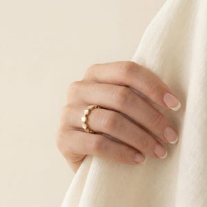 May include: A gold ring featuring a series of small, round, white iridescent stones set along the band. The ring is worn on a finger, showcasing the metallic gold finish and the smooth, circular shape of the white stone inlays.