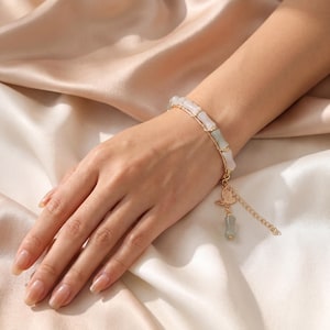 May include: A gold-toned chain bracelet featuring alternating cylindrical white and pale green beads. The bracelet includes a dangling charm shaped like a small leaf and a short extension chain with a single matching pale green bead at the end.