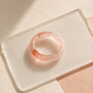 May include: A circular ring featuring a translucent, marbled appearance with blended shades of soft pink, peach, and white. The band has a smooth, polished finish and a consistent thickness throughout its entire circumference.