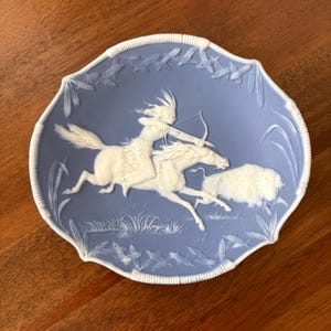 May include: A light blue decorative plate featuring a white relief design of a person riding a horse and aiming a bow and arrow at a buffalo. The plate has a scalloped edge with a white border pattern of leaves and reeds around the perimeter.