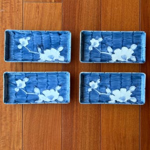 Puede incluir: Cuatro platos rectangulares de cerámica con un fondo azul profundo y un patrón de cuadrícula texturizado. Cada plato muestra un diseño de rama floral blanca con flores abiertas y capullos, dispuestos horizontalmente en el centro.