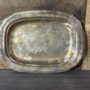 Puede incluir: Una bandeja de servicio ovalada de metal plateado con un borde ancho y liso y un centro hundido. La superficie de la bandeja muestra signos visibles de desgaste, incluyendo arañazos y deslustre, que crean un aspecto metálico envejecido.
